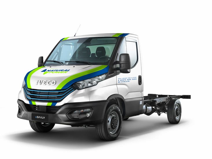 imagem de estúdio do E-Daily, veículo elétrico da IVECO. O veículo aparece por completo mostrando o chassi traseiro. A cabine é na cor prata com listras verde e azul nas laterais.